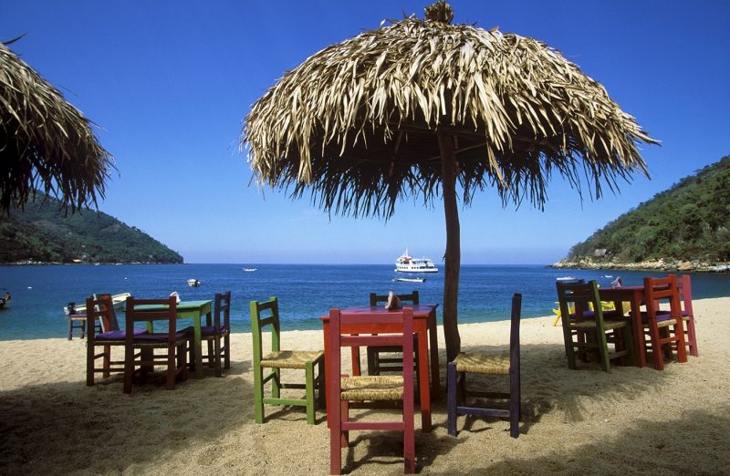 Bay and beach with wooden tables and chairs beneath thatched umbrellas